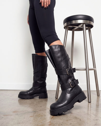 Stivali neri biker con fibbie argento e cinghie incrociate, inquadratura gambe accanto a sgabello