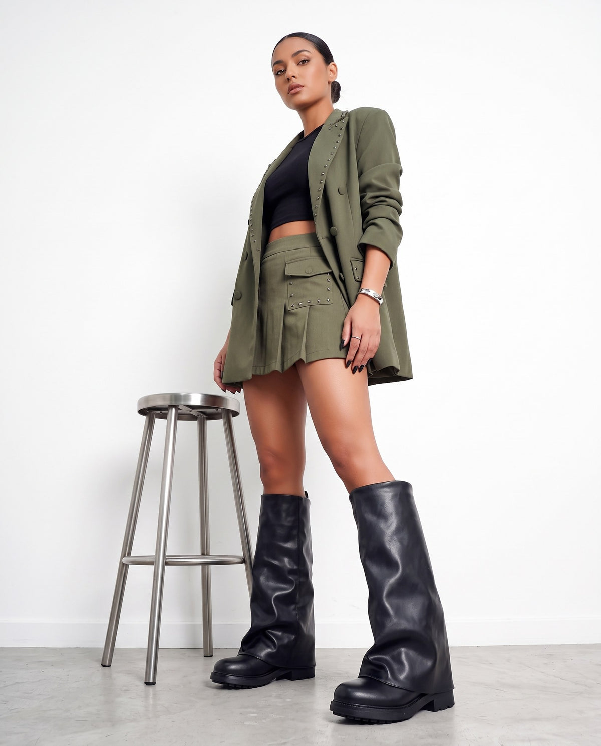 Modella in blazer verde militare e minigonna plissettata, stivali neri a gambale ampio, accanto a sgabello in acciaio