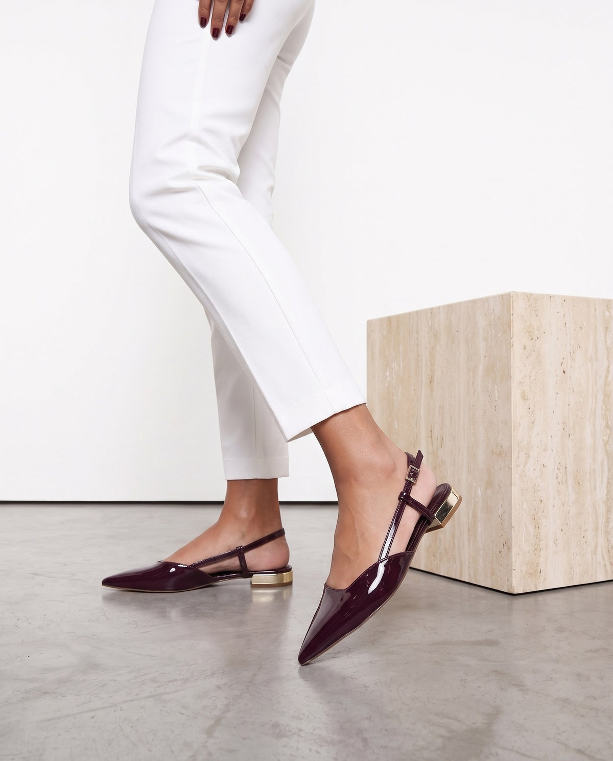 Gambe di modella con pantaloni bianchi e ballerine slingback bordeaux in vernice, tacco gold, vicino a cubo in travertino
