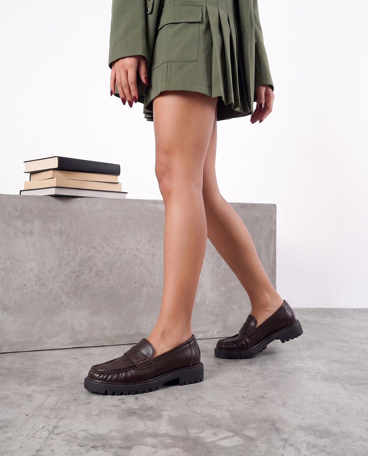 Gambe di modella con mocassini marrone scuro, blazer verde militare, accanto a blocco di cemento con libri