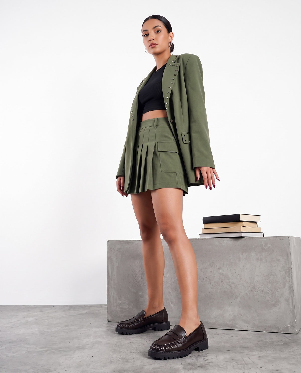 Modella in blazer verde militare e minigonma plissettata, mocassini marroni, accanto a blocco di cemento con libri