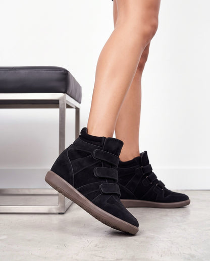 Primo piano di gambe con sneaker wedge nere a tre strappi, suola marrone, accanto a una panca cromata con seduta in pelle nera