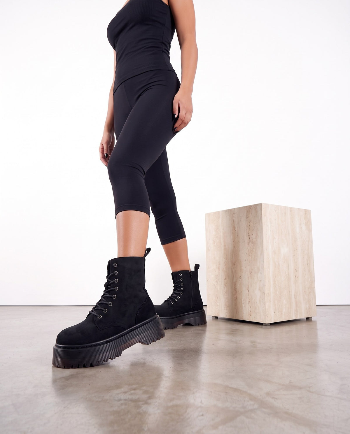 Gambe di modella con stivaletti neri scamosciati con platform, vicino a un cubo di travertino, su pavimento in cemento