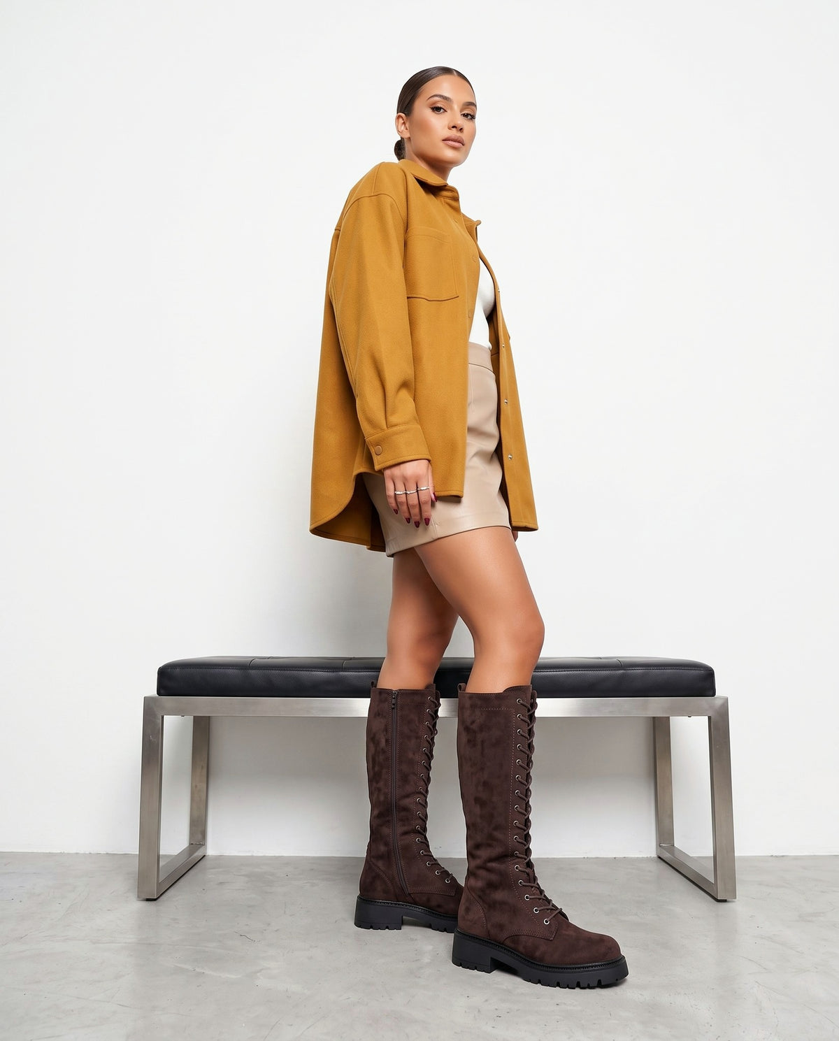 Modella in piedi con stivali alti in camoscio marrone, giacca senape e minigonna beige, panca sullo sfondo.