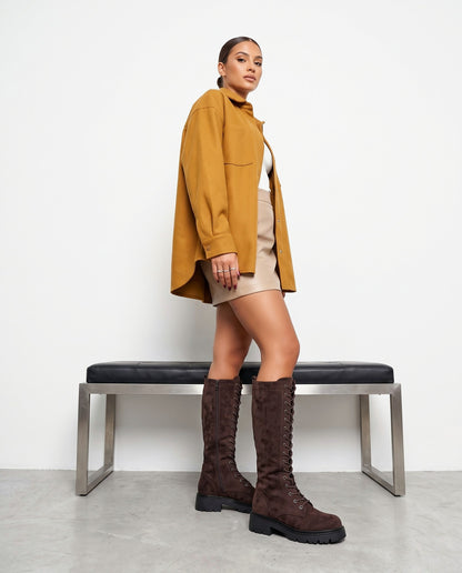 Modella in piedi con stivali alti in camoscio marrone, giacca senape e minigonna beige, panca sullo sfondo.