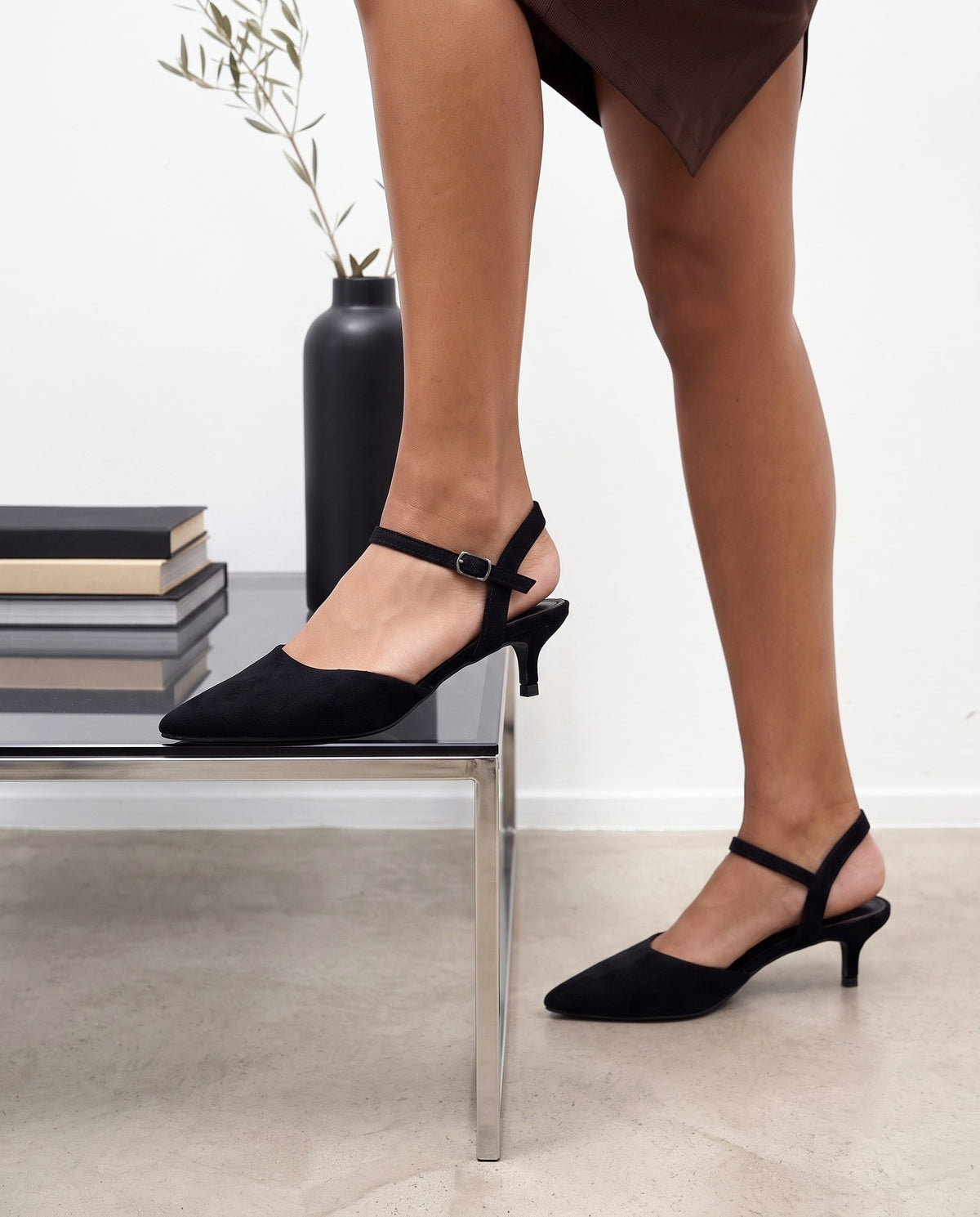 Dettaglio ravvicinato di scarpe décolleté nere in camoscio con cinturino, su sfondo con tavolino e vaso nero