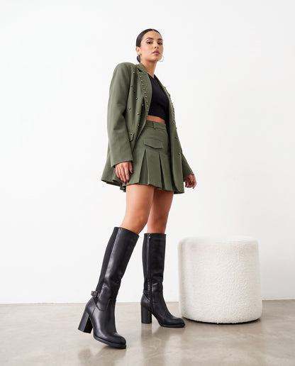 Modella in piedi con blazer verde militare, minigonna plissettata e stivali neri al ginocchio, accanto a pouf bianco.