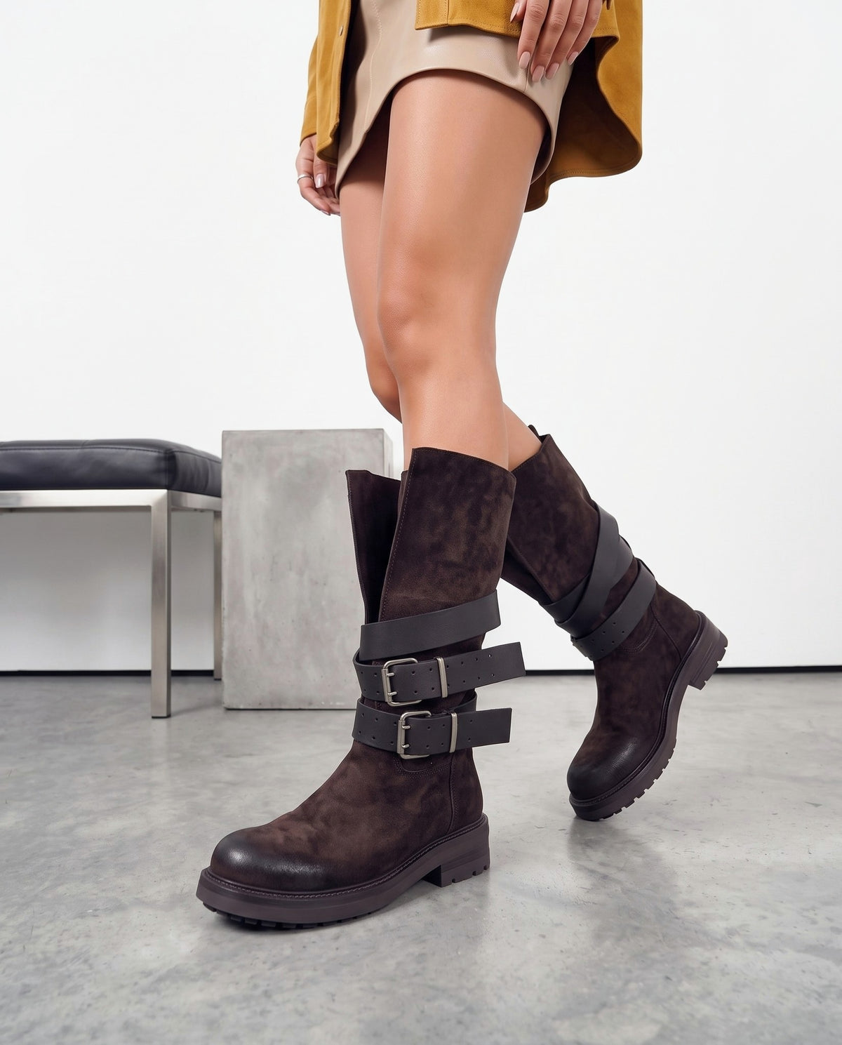 Gambe femminili con stivali alti in camoscio marrone scuro con doppia fibbia, su pavimento in cemento chiaro
