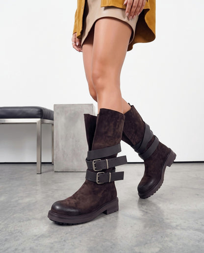 Gambe femminili con stivali alti in camoscio marrone scuro con doppia fibbia, su pavimento in cemento chiaro