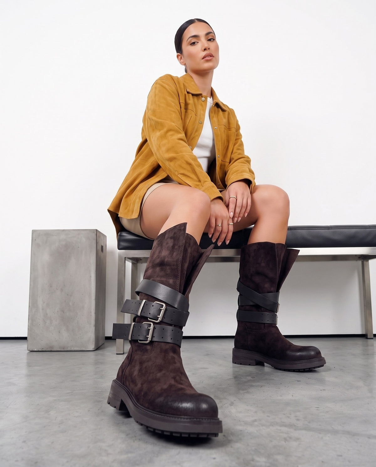 Modella seduta con stivali marrone scuro a cinturini neri, gambe accavallate, su set con panca e blocco in cemento