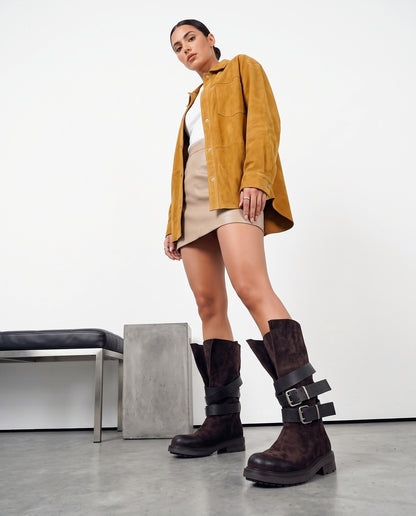 Modella in piedi con stivali marroni in camoscio a doppia fibbia, giacca senape e minigonna beige