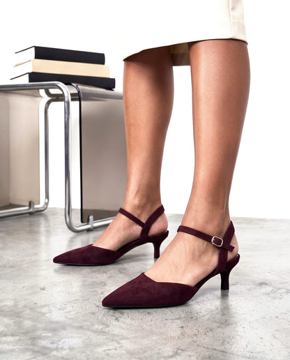 Primo piano di due scarpe décolleté bordeaux in camoscio con cinturino alla caviglia, tacco basso, su pavimento in cemento