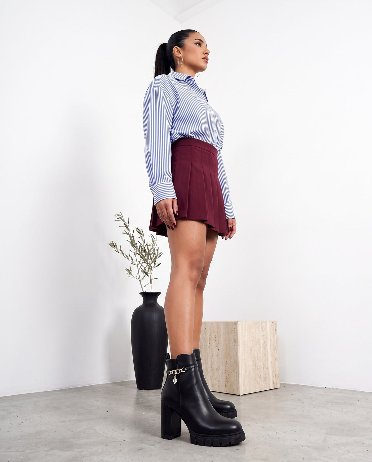 Modella in piedi di profilo con stivaletti neri, camicia a righe e minigonna bordeaux, in studio minimal.