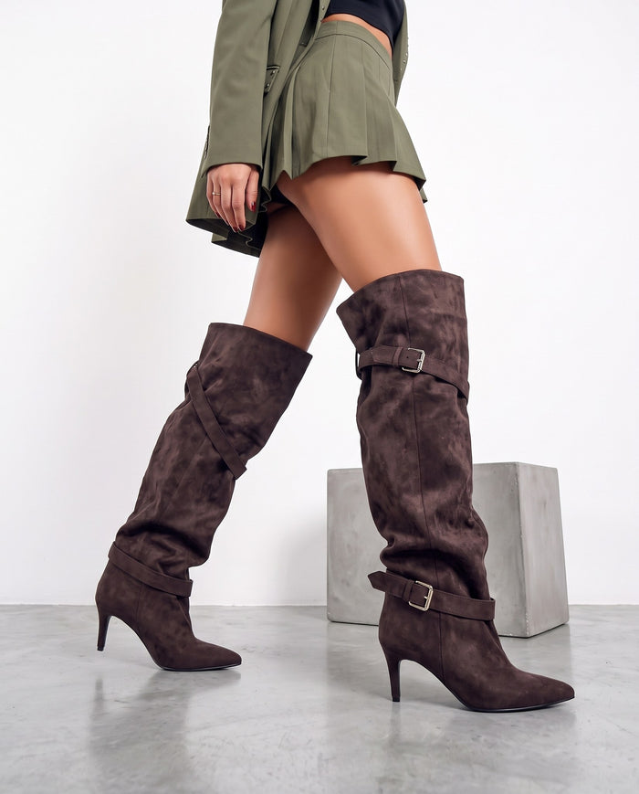 Gambe femminili con stivali alti in camoscio marrone, doppie fibbie, tacco a spillo, su pavimento in cemento chiaro