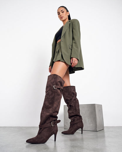 Modella in piedi di profilo con stivali marroni scamosciati sopra il ginocchio, blazer verde militare e minigonna plissettata.