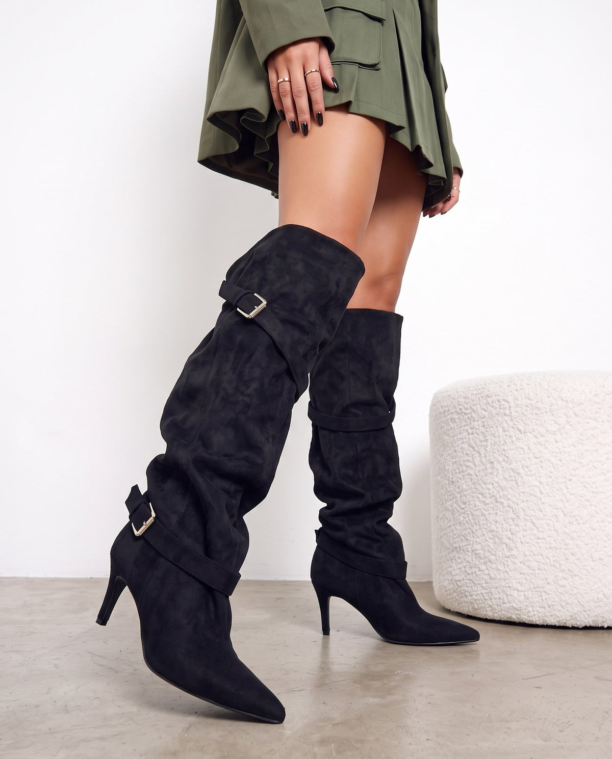 Gambe di modella con stivali alti in camoscio nero con fibbie, accanto a pouf bianco bouclé