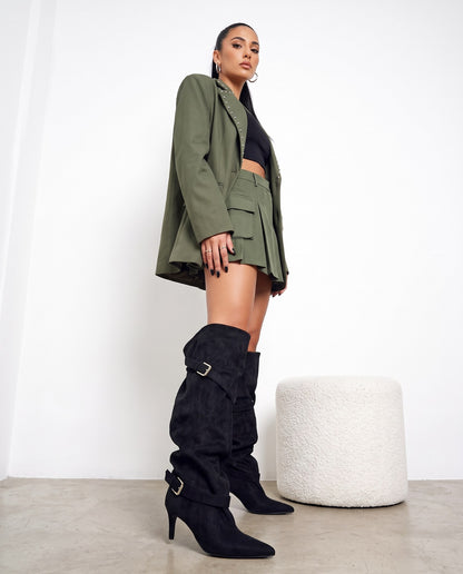 Modella di profilo con blazer verde militare, minigonna e stivali alti neri scamosciati con fibbie