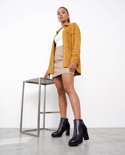 Modella in piedi con stivaletti neri, giacca senape, gonna beige, accanto a sgabello in acciaio.