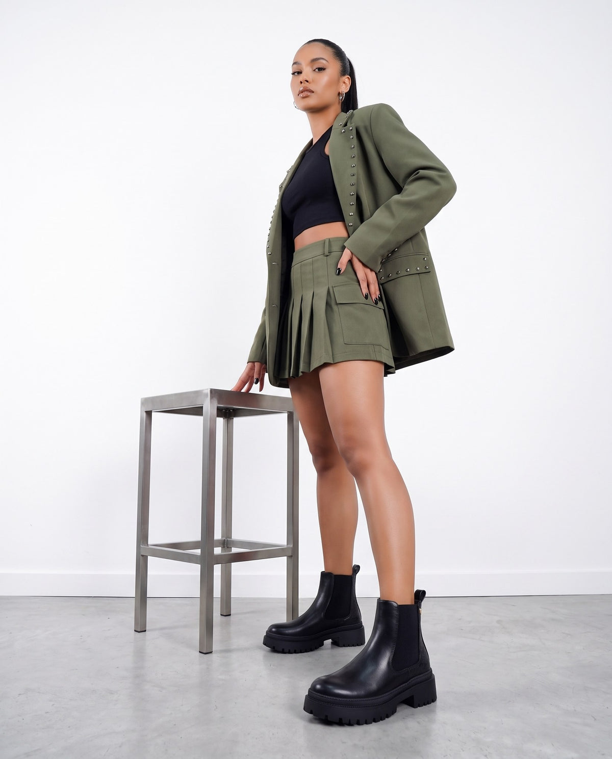 Modella in piedi con blazer verde militare, minigonna plissettata e stivaletti neri chunky, accanto a uno sgabello in acciaio.