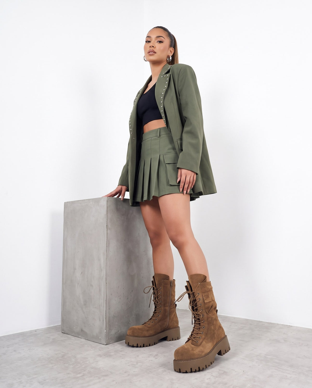 Modella in blazer e minigonna verde militare con stivali camoscio marrone, accanto a blocco di cemento