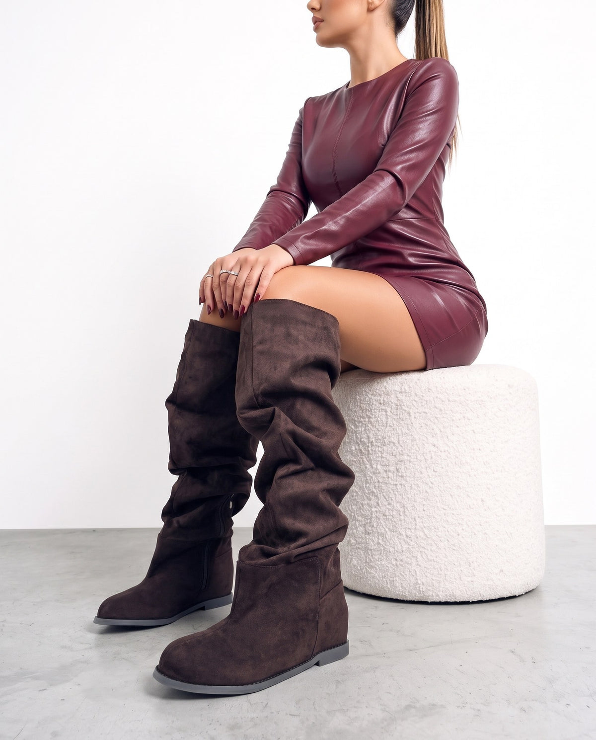 Modella seduta su pouf bianco, indossa stivali alti in camoscio marrone scuro, gambe accavallate.