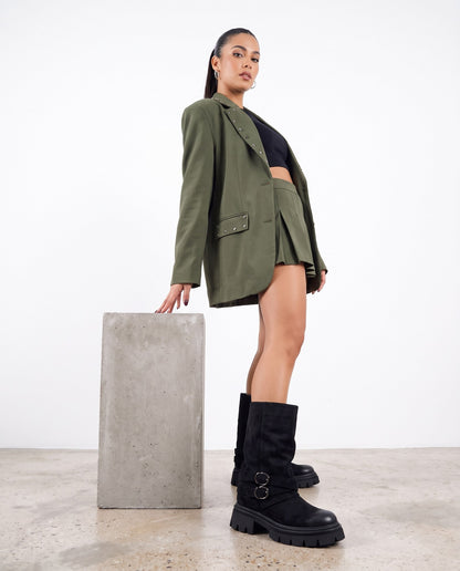 Modella in blazer e minigonna verde militare con stivali neri camoscio, accanto a blocco di cemento