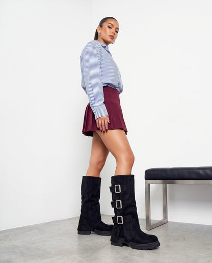 Modella in piedi di profilo con stivali neri scamosciati con tre fibbie, camicia a righe e minigonna bordeaux