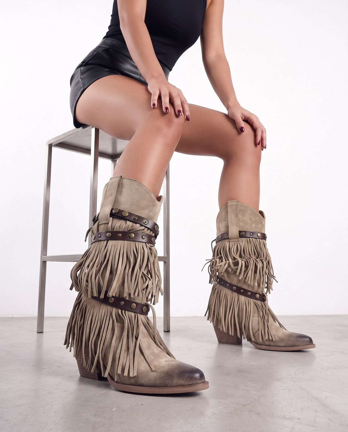 Modella seduta su sgabello, gambe incrociate, in primo piano stivali beige con frange e borchie