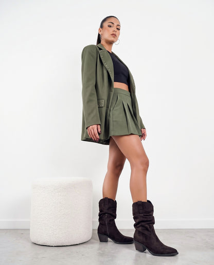Modella in piedi con stivali western marroni in camoscio, blazer e minigonna verde militare, pouf bianco accanto.