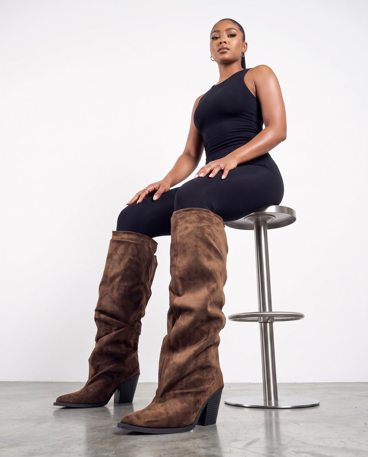 Donna seduta su sgabello cromato con stivali alti in camoscio marrone, mani sulle ginocchia con anelli