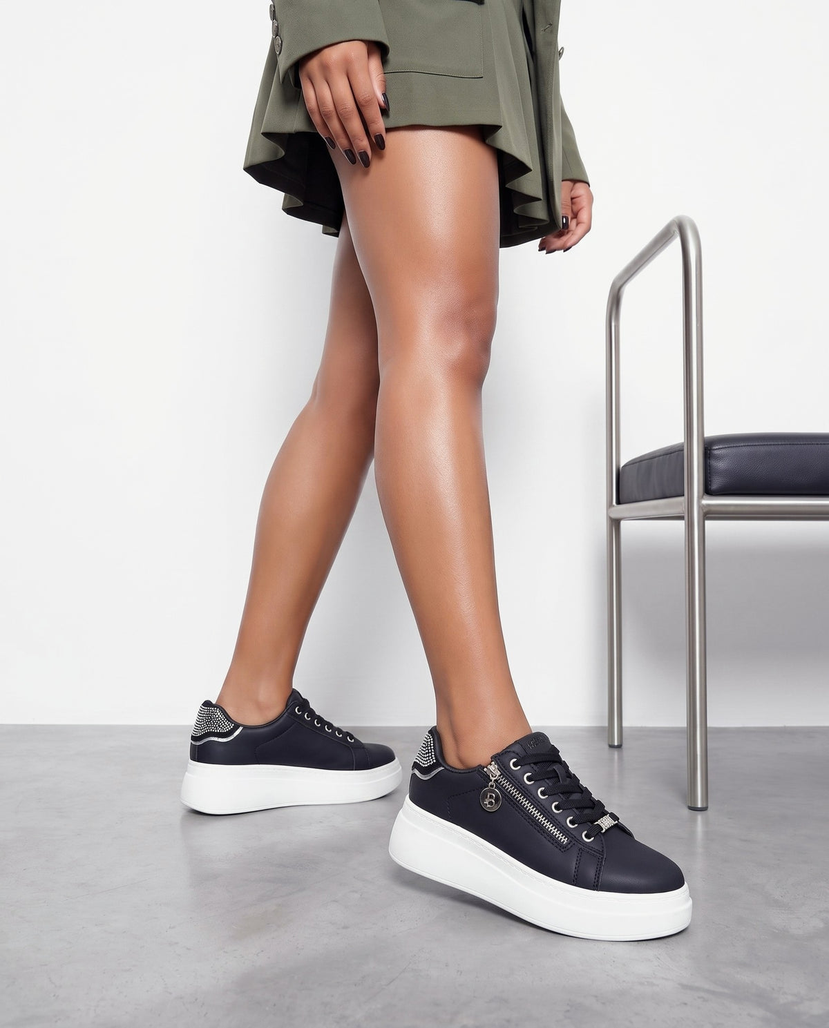 Gambe della modella con sneakers nere platform zip e strass, sgabello in acciaio accanto, pavimento in cemento