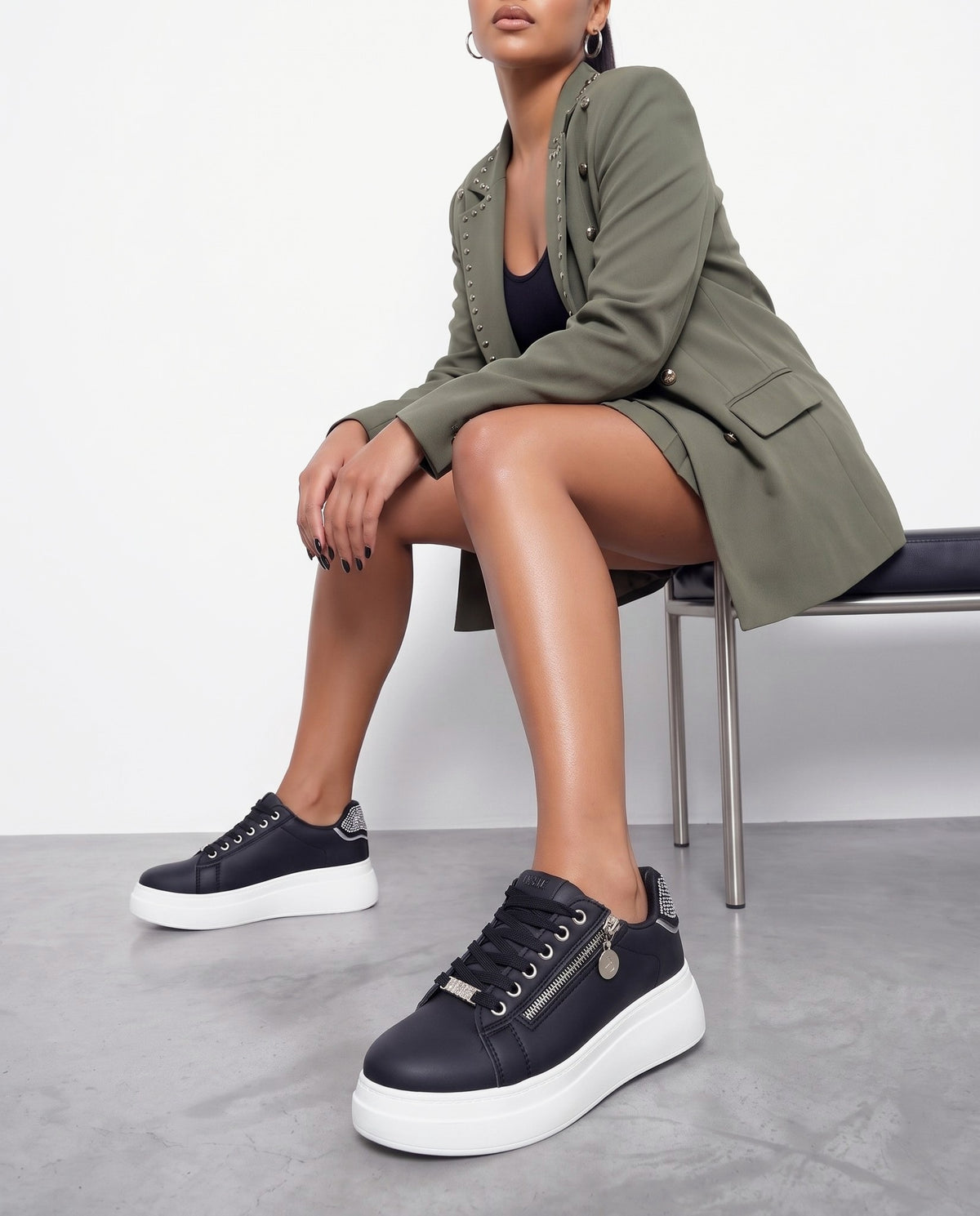 Modella seduta su panca, indossa sneaker nere con zip e strass, gonna e blazer verde militare