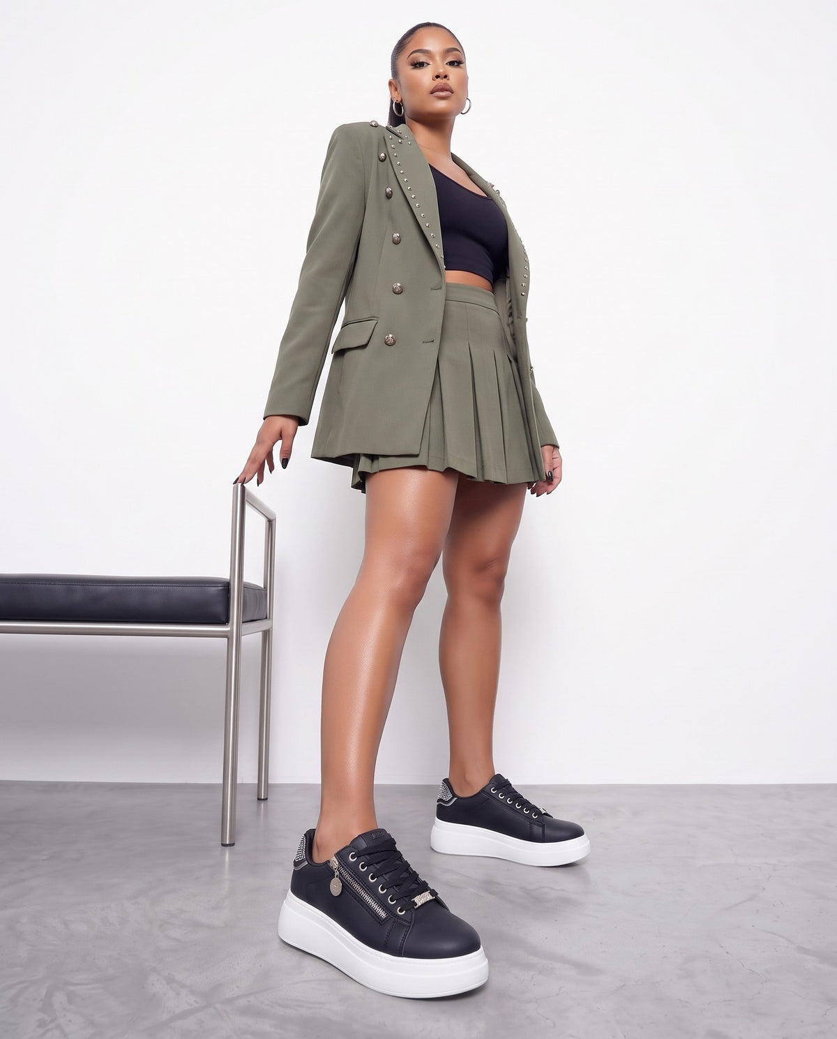 Modella in blazer e minigonna verde militare che cammina verso la camera con sneakers nere e bianche.